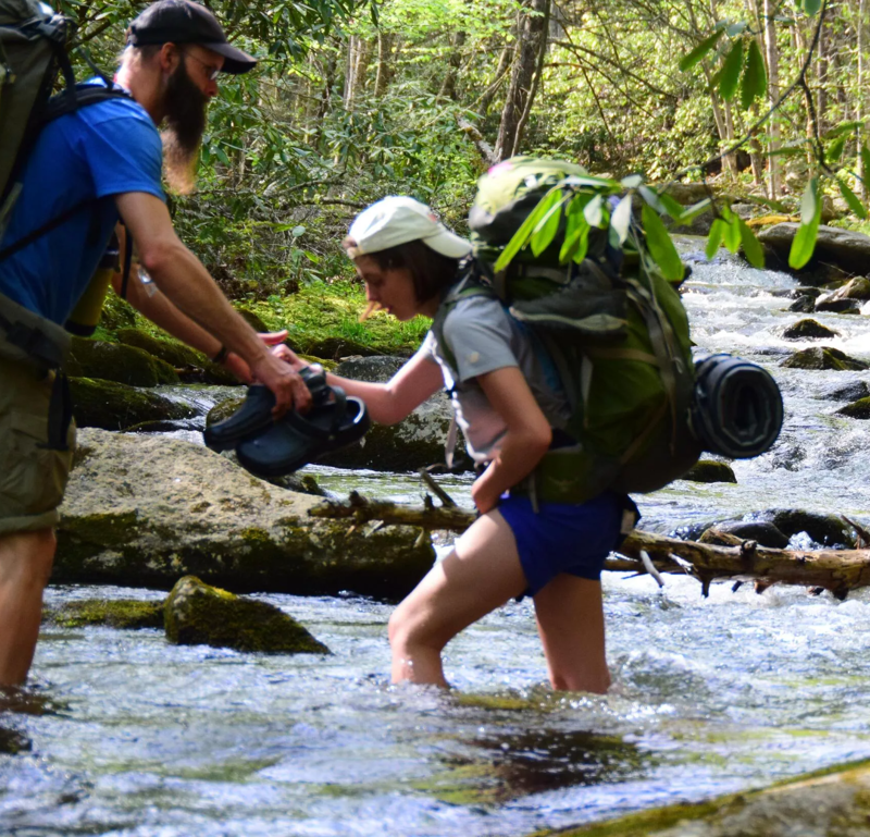 In the image, a man with a beard is assisting a younger person, possibly a teenager, as they cross a shallow stream. Both are wearing backpacks, suggesting they are hiking or backpacking. The man is handing the younger person a pair of shoes, presumably to wear while crossing the water. The stream is surrounded by rocks and trees, indicating a natural, outdoor setting.